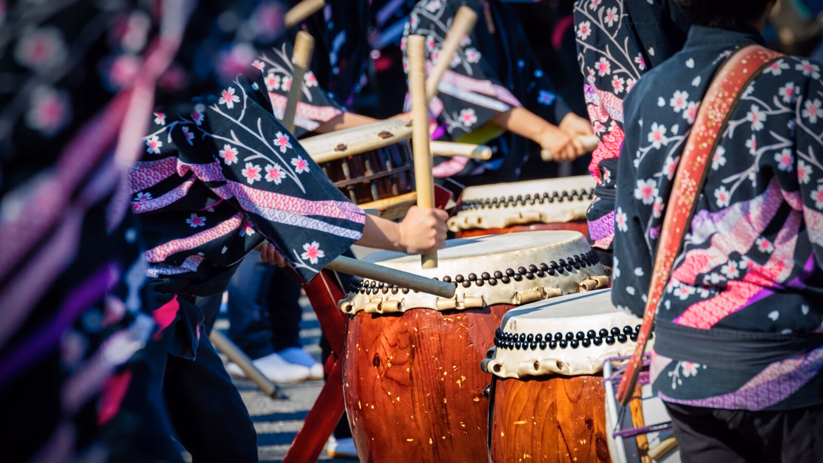 Close up of people hitting large wooden drums with thick wooden sticks in a ceremonial performance. The drummers are wearing traditional robes decorated with cherry blossom patterns.