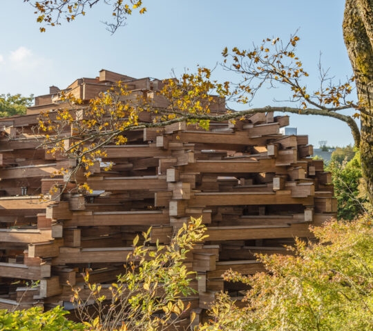 Large circular structure made of stacked wooden beams in a grassy park with trees.