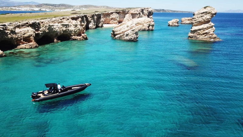 A black RIB boat navigating crystal clear turquoise water by rocky island cliffs.