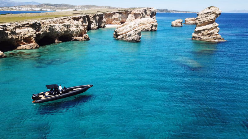 A black RIB boat navigating crystal clear turquoise water by rocky island cliffs.