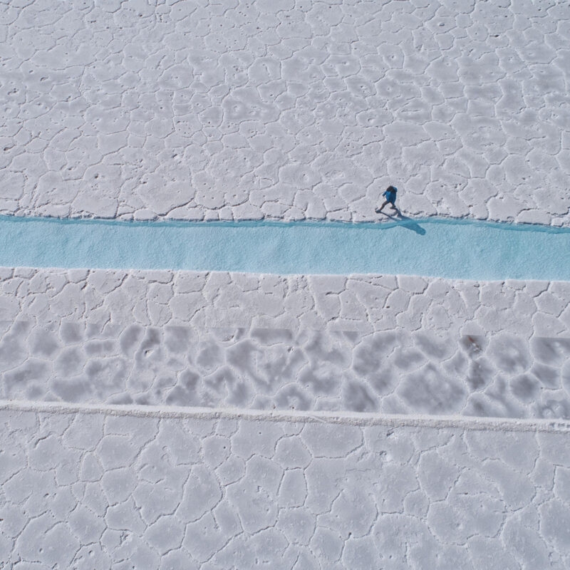 Overhead view of a person in blue walking on a strip of blue water across a vast white textured salt flat. Luxury Argentina tours.
