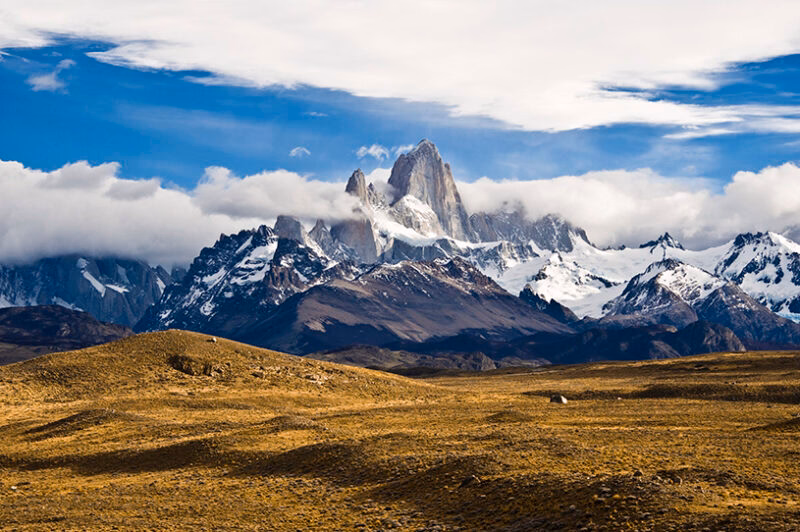 The jagged, snow-capped Mount Fitz Roy rises above a golden grassy foreground under a blue, cloudy sky. Luxury Argentina holidays.