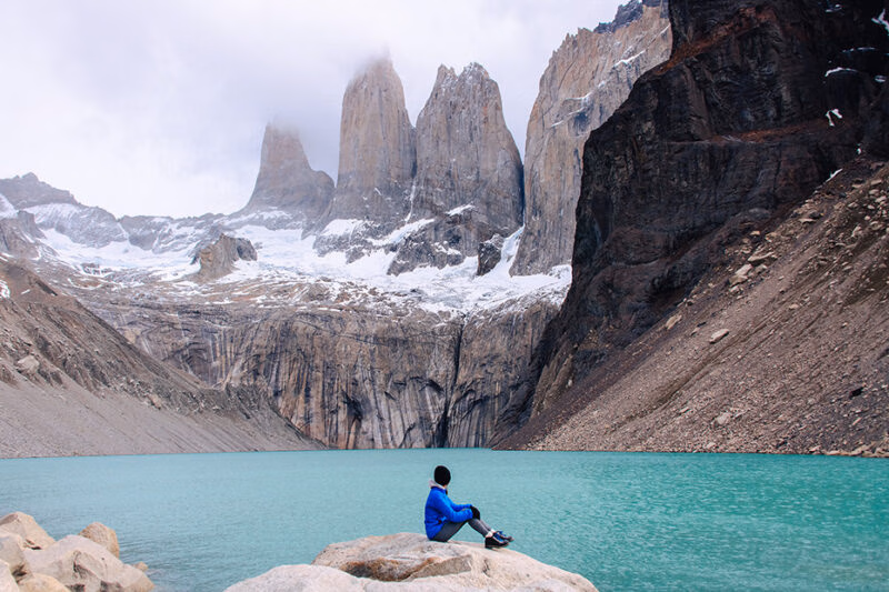 A person in a blue jacket looking at large rock towers and a blue lake on luxury Chile vacations.