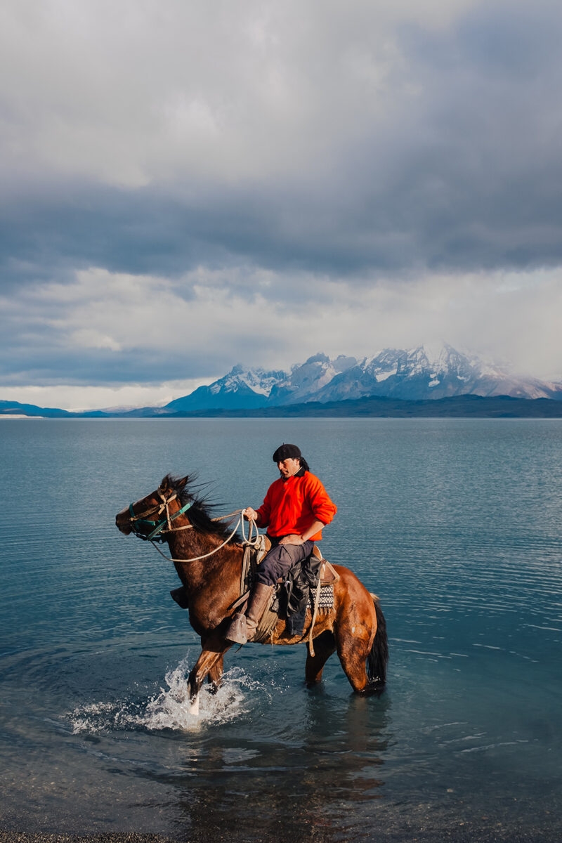 A man riding a horse through blue water with mountains behind him on luxury Chile trips.