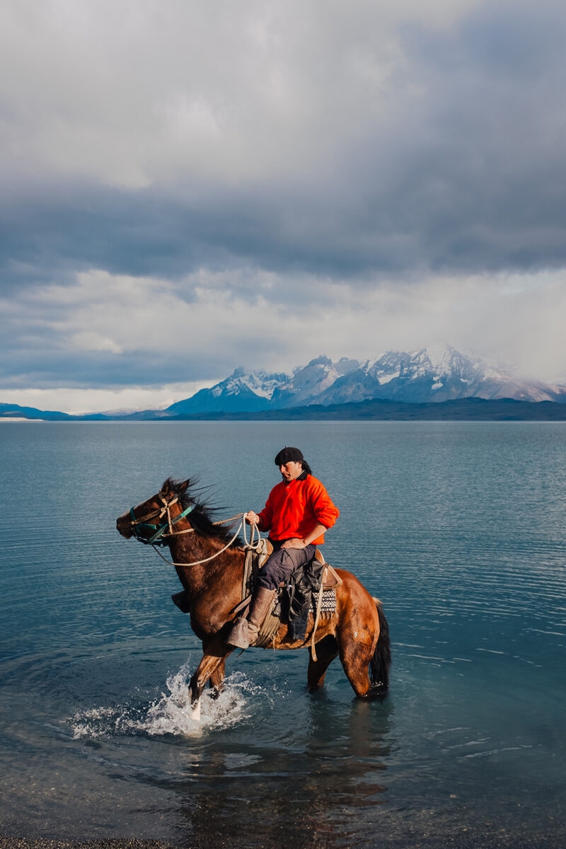 A man riding a horse through blue water with mountains behind him on luxury Chile trips.