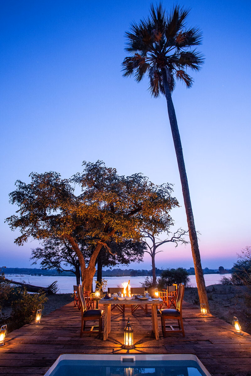 A wooden deck set for a dinner, lit by lanterns under a tall palm tree with a river and sunset in the background.