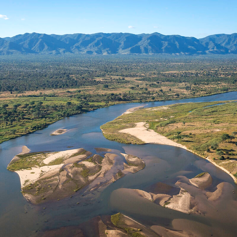Aerial view of a wide river with sandbanks running through a forested valley with a large range of mountains in the distance.