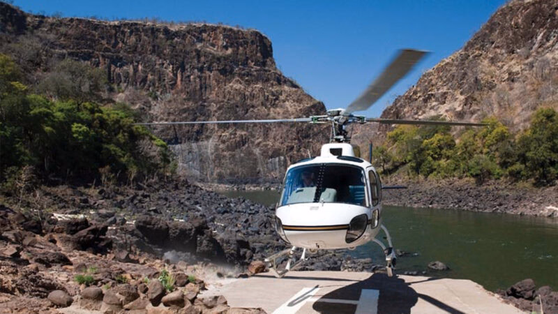 A white helicopter lifting off a landing pad on a rocky bank next to a river and steep, rocky cliffs.