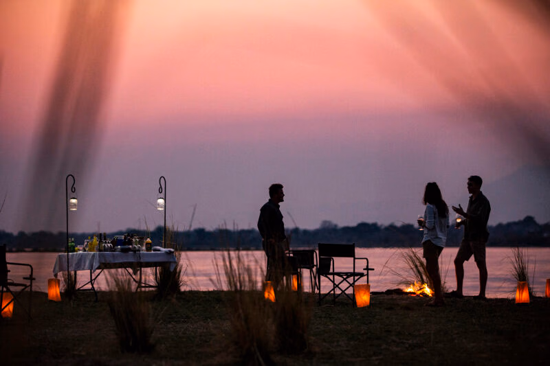 Silhouettes of three people standing by a river at sunset, with a bar table and lanterns lit on the grassy bank.