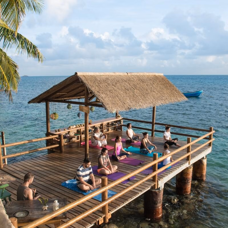 A group of people practice yoga and meditation in a seated position on mats on a wooden deck with a thatched roof, extending out over the turquoise ocean during Luxury Colombia Holidays.