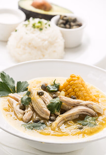A close-up of a bowl of Ajiaco, a creamy chicken and potato soup with capers and corn, served with rice and avocado in the background during Luxury Colombia Trips.