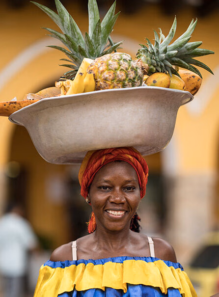 Portrait of a smiling woman in a colorful yellow and blue dress with a red and orange turban, balancing a basin of fruit on her head during Luxury Colombia Holidays.