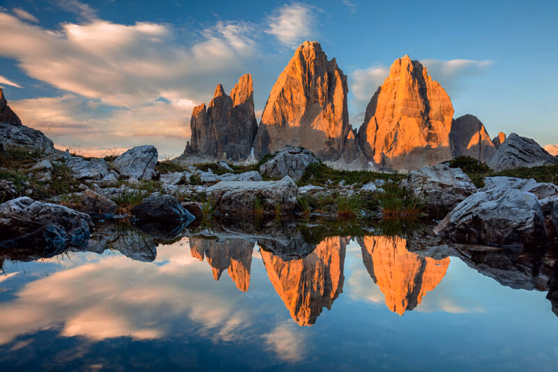 Luxury Italy Tours Three jagged mountain peaks at sunset reflected in the clear water of a small mountain lake.