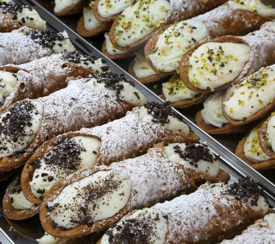 Luxury Italy Tours - Rows of typical Sicilian cannoli, a crisp pastry cylinder stuffed with ricotta cheese and sprinkled with powered sugar, nuts, and chocolate shavings