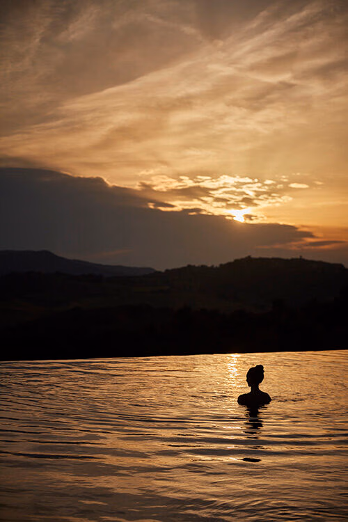 Luxury Italy Tours - The silhouette of the head a shoulders of a woman with long hair piled on top of her head in a bun style, sitting in calm water overlooking hills and mountains during a cloudy, golden sunset