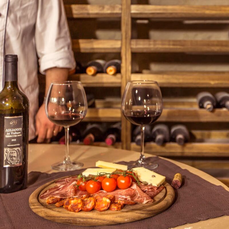 Luxury Italy Tours - A wooden round plate on a table laden with fresh small tomatoes, cured meats, and cheese. There are also two wine glasses with a small amount of red wine, and a green wine bottle with the cork laid next tot he plate. In the background, there are rows of wine bottles laying on wooden shelves in a cellar, and we see the arm of a person wearing a chef's white jacket