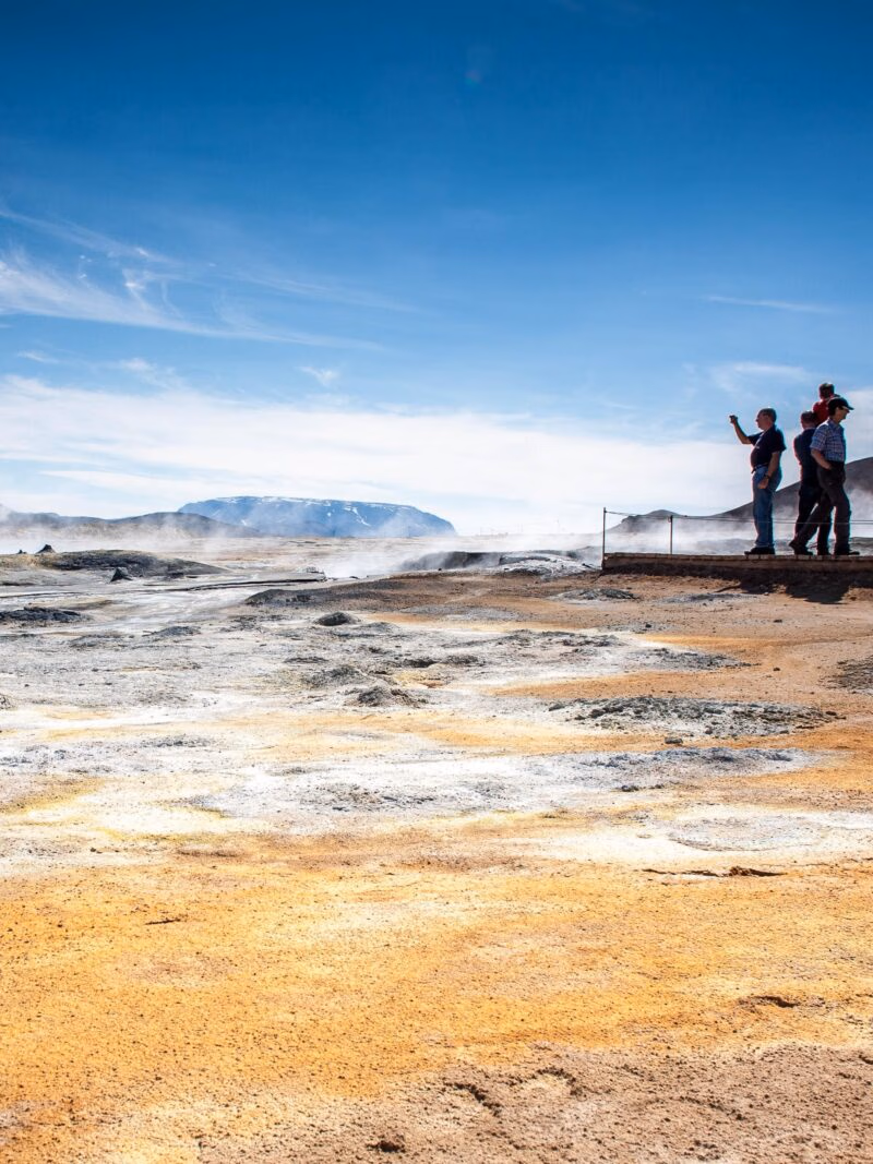 People view a steaming, rocky geothermal landscape with yellow and white mineral deposits under a clear blue sky.