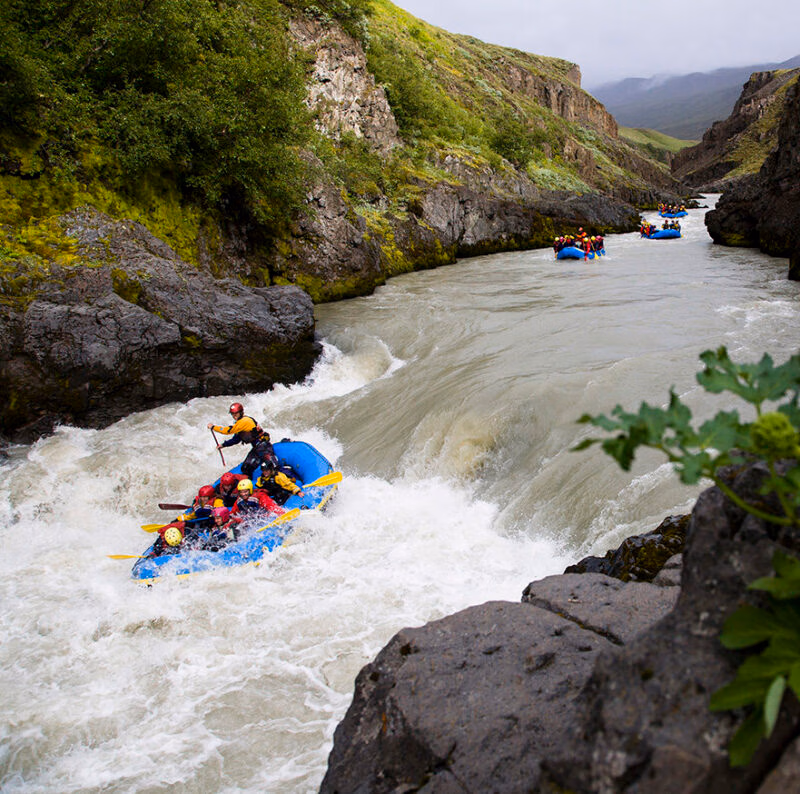 Adventurers in blue rafts paddle through white water rapids in a deep canyon surrounded by green cliffs.