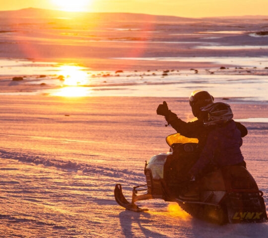 Silhouette of two people on a snowmobile at sunset, with one person giving a thumbs up.