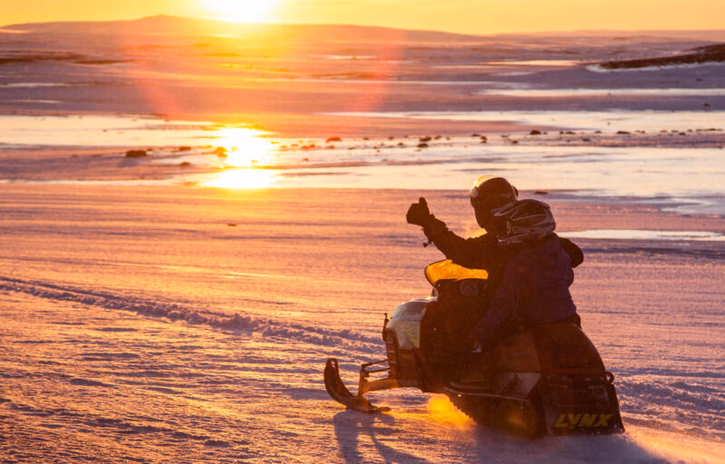 Silhouette of two people on a snowmobile at sunset, with one person giving a thumbs up.
