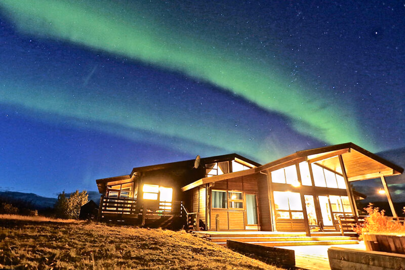 A modern wooden cabin glows at night under the bright green Aurora Borealis, an ideal setting for luxury Iceland holidays.