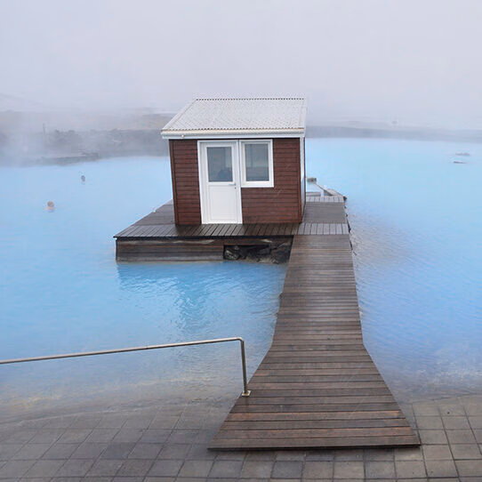 A small brown wooden cabin on a deck is surrounded by the milky blue water of the Blue Lagoon, promoting luxury Iceland tours.