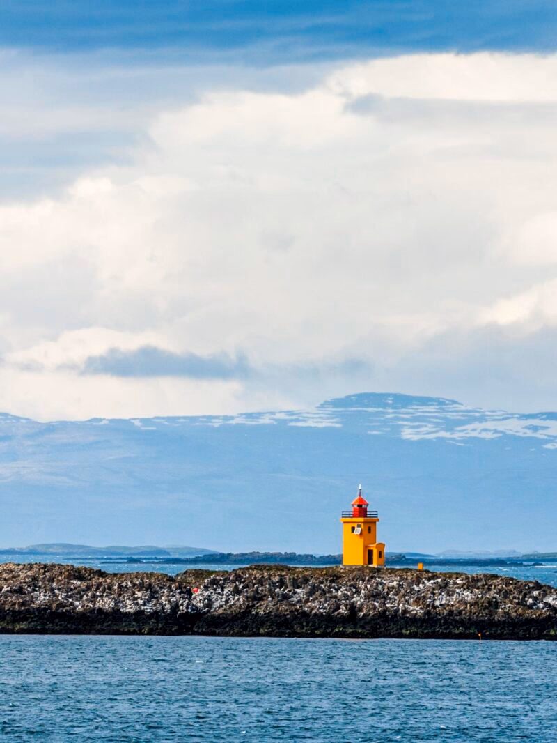 A small, bright yellow lighthouse stands on a dark rocky breakwater