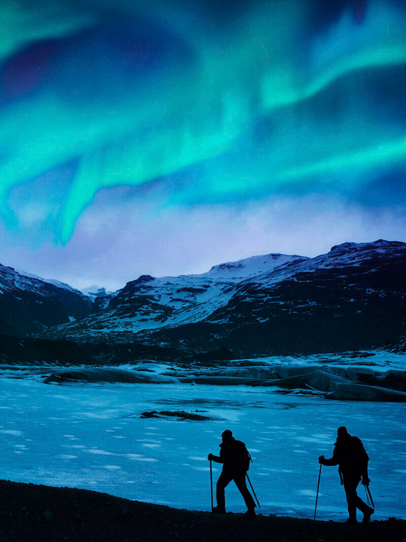 Two hikers stand silhouetted on a frozen glacier under the blue and green Northern Lights, highlighting luxury Iceland tours.