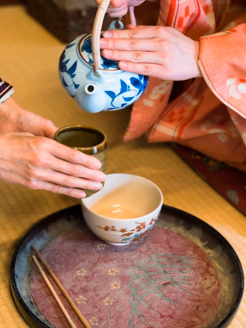 Private Japan Luxury Tours - detail of two women in traditional kimono, kneeling on tatami preparing, pouring a cup of tea which is in the hands of one woman. They are in traditional Japanese old house on tatami.