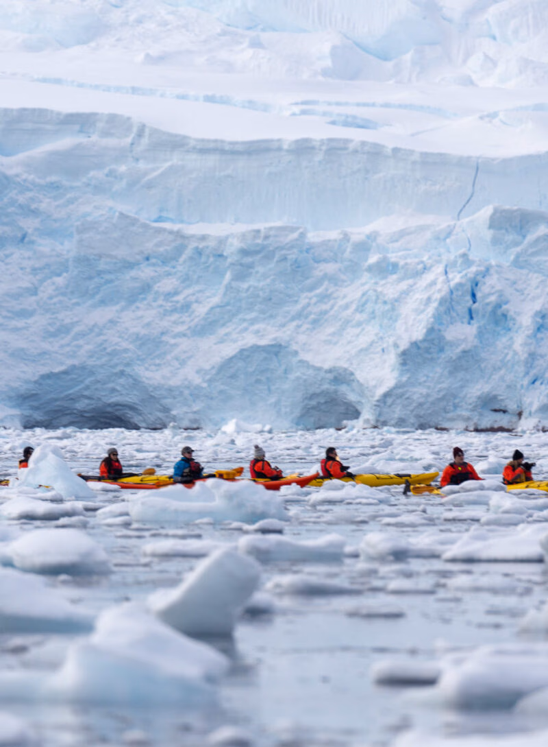 Several people in yellow kayaks are paddling near a massive blue and white glacier wall with floating ice chunks.
