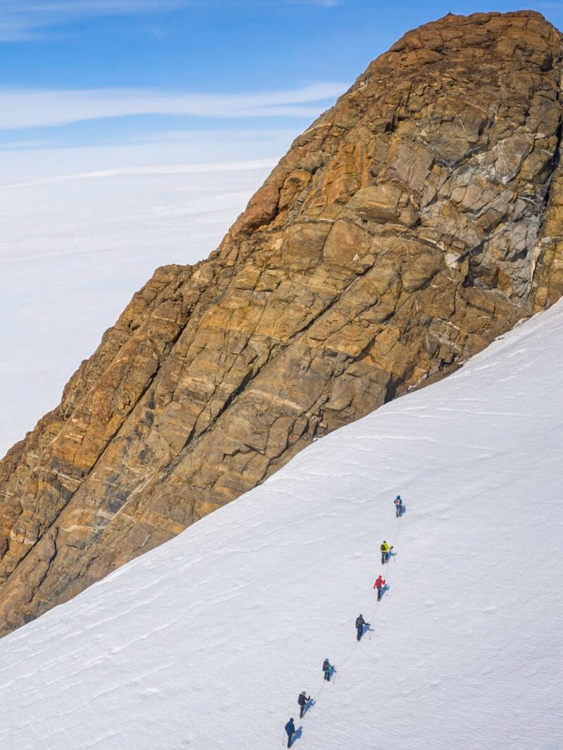 A line of climbers ascend a steep snow and ice slope next to a massive, brown, rocky mountain wall.