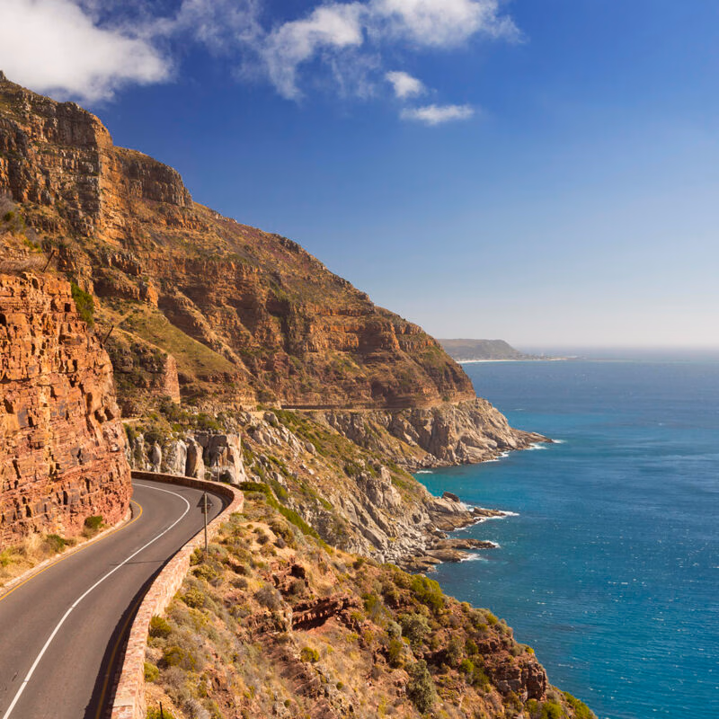 Luxury South African Tours - A high-angle view of a coastal road curving along steep mountains next to the blue sea.