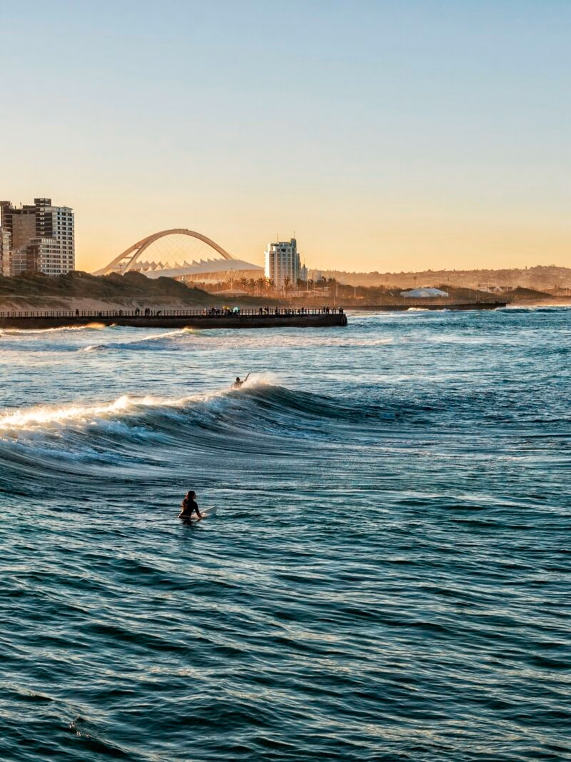 Luxury South African Tours - Person on a surfboard in the water with a pier and a distinctive arched stadium in the background.