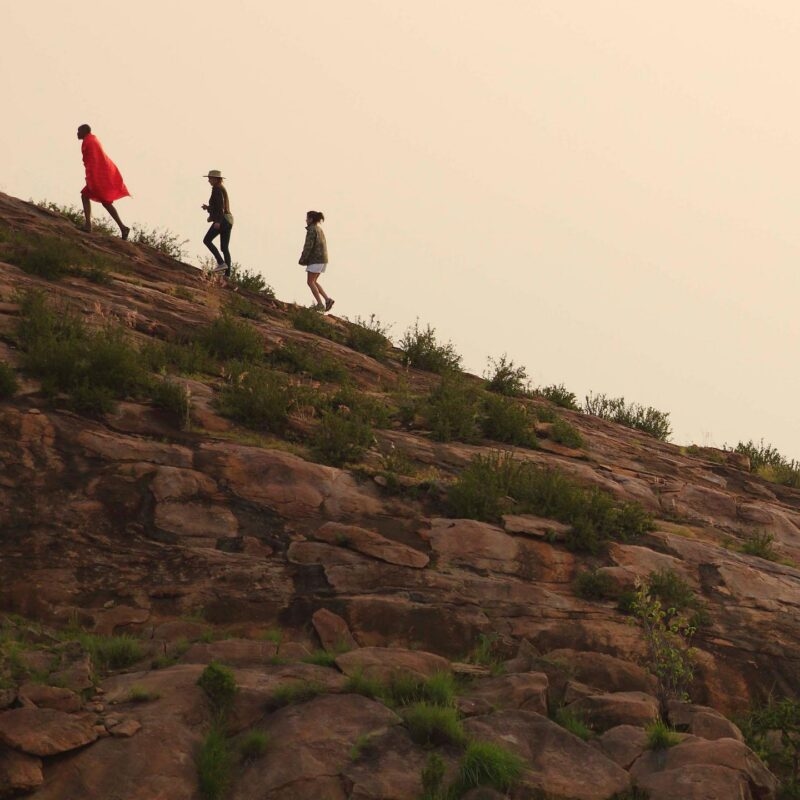 A group on a luxury Kenya family safari hikes up a rocky slope following a guide in a red cloak.
