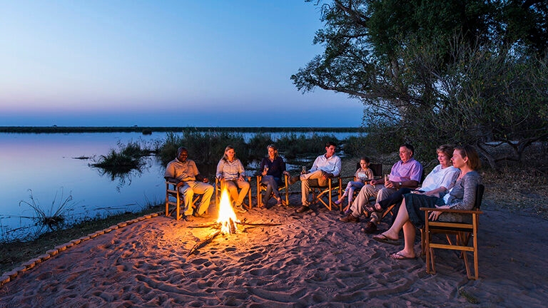 People sitting around a campfire at dusk next to a lake or river.