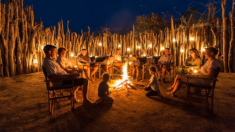 People sitting in a circle around a campfire surrounded by a tall wooden fence at night.