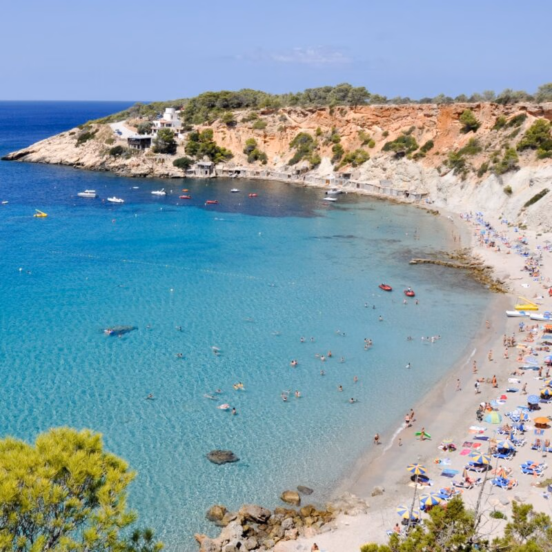 Aerial view of a crowded beach on a turquoise bay backed by a red cliff. A perfect location for luxury Spain trips.