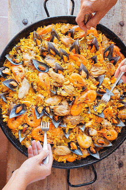 Overhead view of a large pan of seafood paella with several hands holding forks, representing luxury Spain tours.