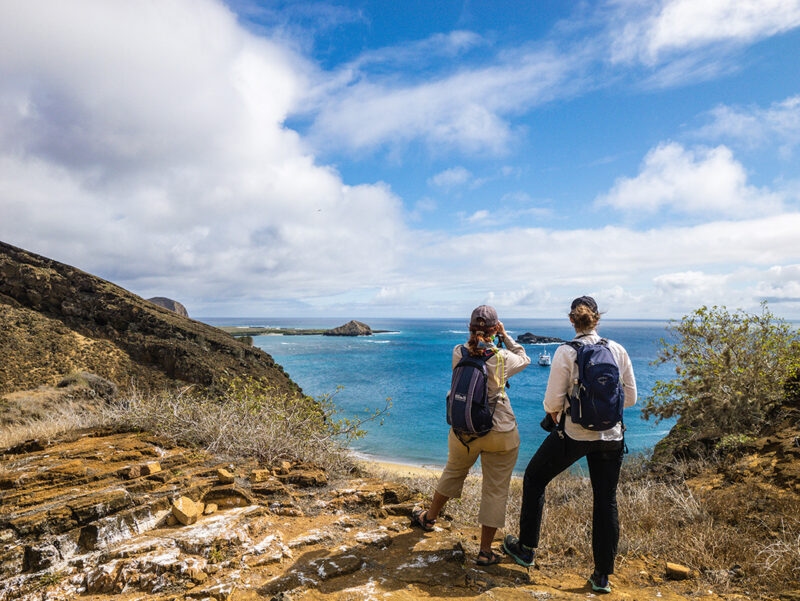 Two people with backpacks look out from a rocky bluff over a bay and distant volcanic islands. luxury Galapagos vacations.