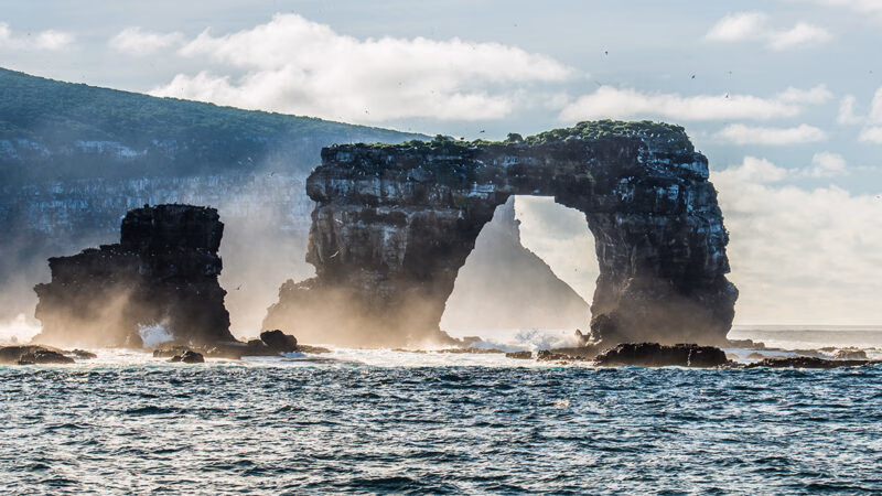 Darwin's Arch, a large natural rock arch formation rising from the ocean, waves crashing at its base. luxury Galapagos cruises.