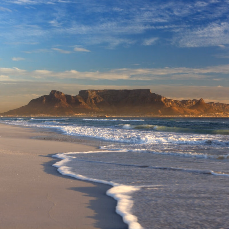 Table Mountain looms over a sandy beach with white sea foam washing ashore under a blue and cloudy sky.