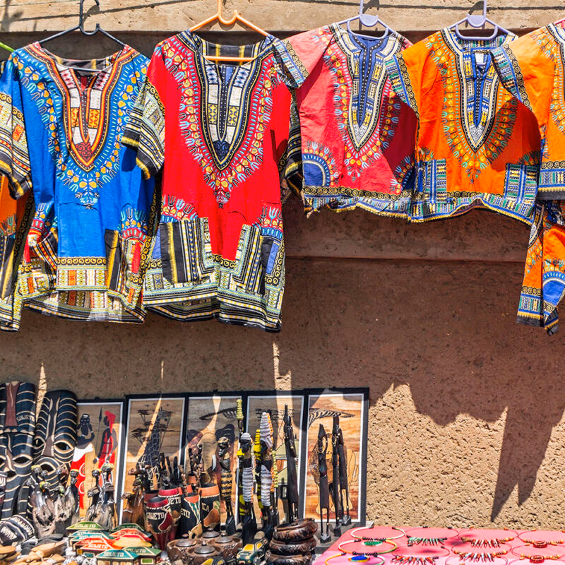 Brightly colored dashiki shirts and various handcrafted African souvenirs displayed for sale at an outdoor market stall.