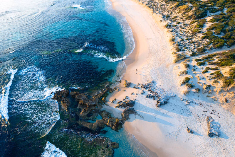 Aerial view of a rugged, curved coastline with white sand, turquoise water, and breaking waves, a beautiful stop on luxury Australia trips.