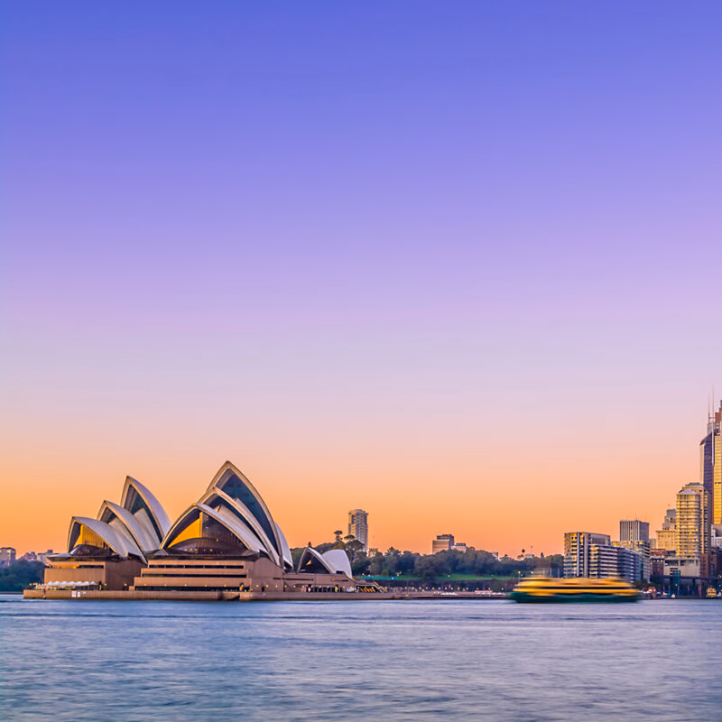The Sydney Opera House stands by the water at sunset with a cityscape in the background and a purple-orange gradient sky, on luxury Australia tours.