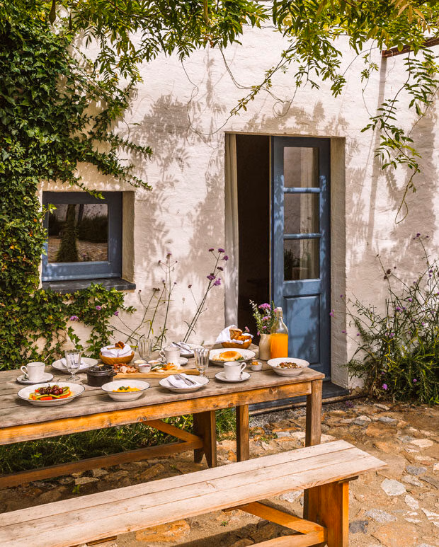 Outdoor dining table set for breakfast in a sunny courtyard next to a white building with a blue door, ideal for luxury Portugal vacations.