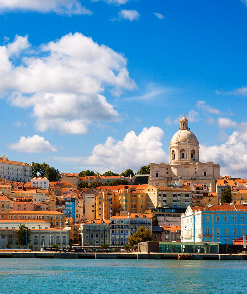 View of Lisbon waterfront with colorful buildings and a large dome church (Santa Engrácia) under a blue sky, luxury Portugal vacations.