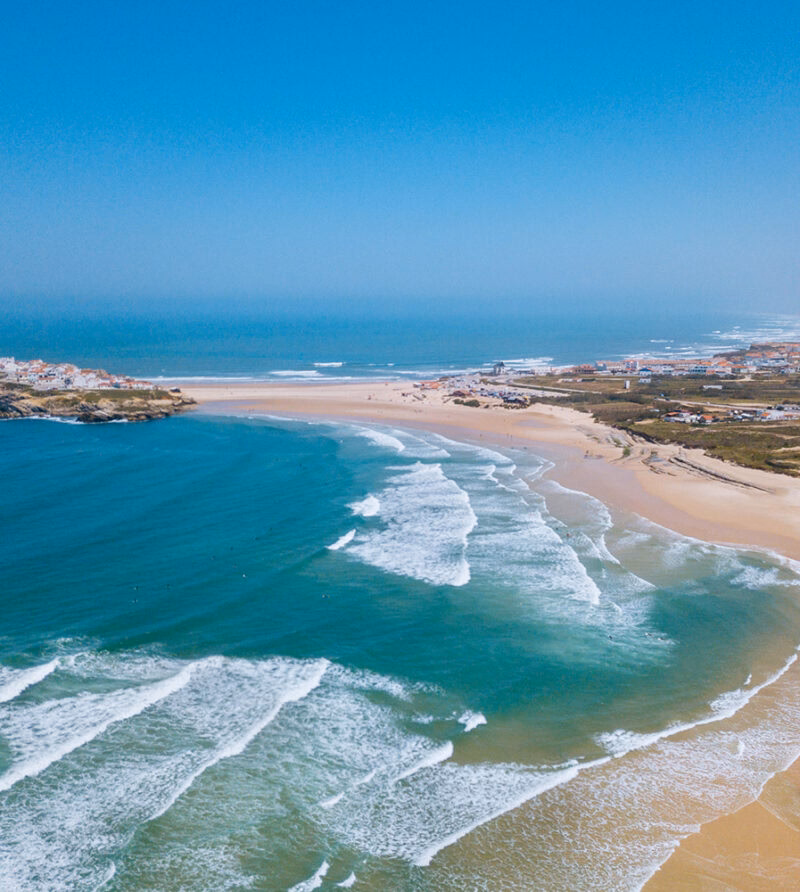 Aerial view of a wide sandy beach, turquoise ocean waves, and a coastal village under a clear sky, ideal for luxury Portugal vacations.