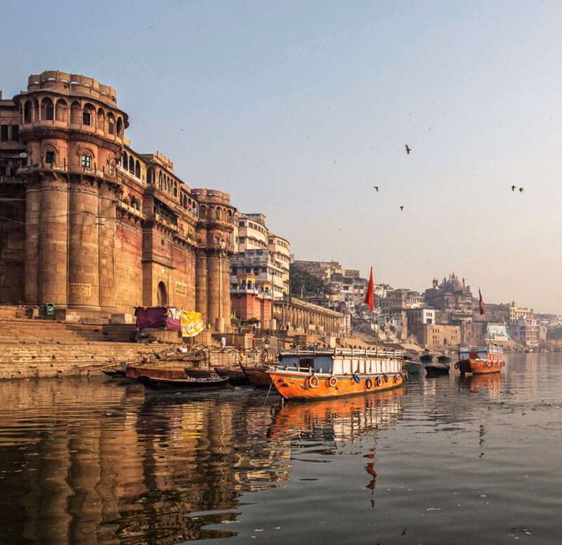 Traditional stone ghats and buildings line the Ganges River, with several boats docked in the foreground in Varanasi on luxury India tours.