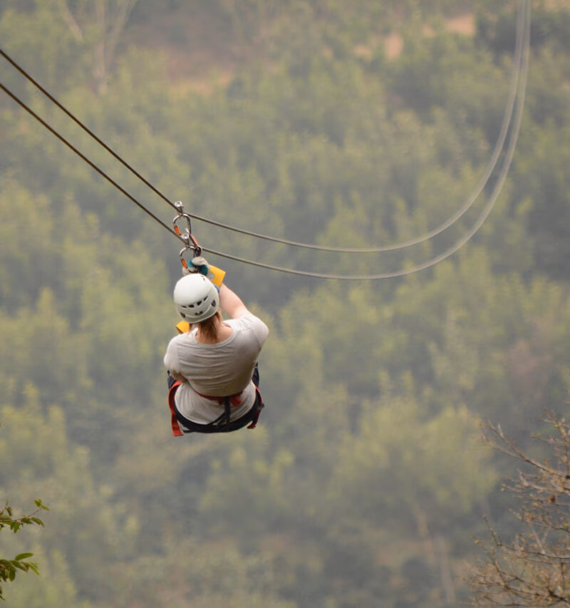A person in a white helmet and shirt zip-lining high over a dense, slightly hazy green forest landscape.