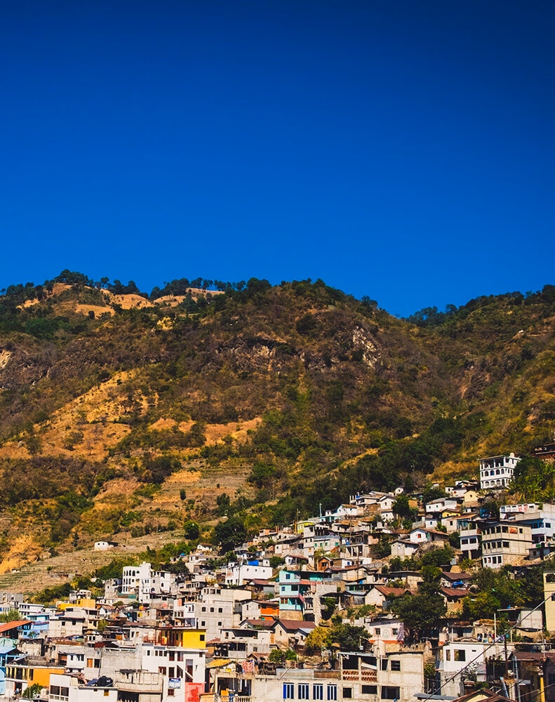 Village houses densely packed on a steep, dry hillside with green trees at the top, under a clear, bright blue sky.